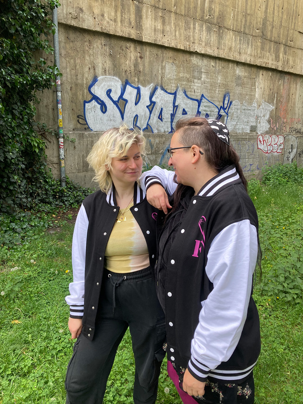 Auf dem Foto stehen Riv Elinson und Nui Arendt nebeneinander und lächeln sich an. Nui hat den rechten Arm auf Rivs Schulter gelegt. Beide tragen eine Schwarz-weiße College-Jacke. Nui trägt eine Kippa mit einem Schnurrbart-Muster auf dem Kopf.